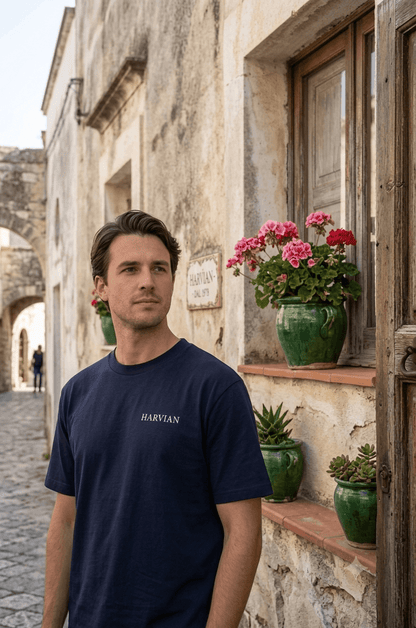 Man wearing HARVIAN Capri Tee standing near flower pots on a rustic street.